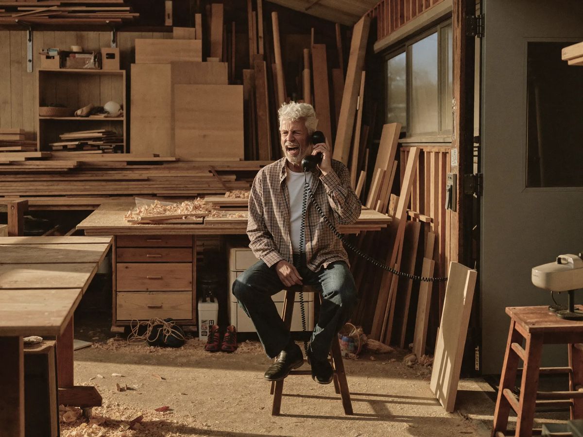 A carpenter in his sixties laughing on a phone call in his workshop